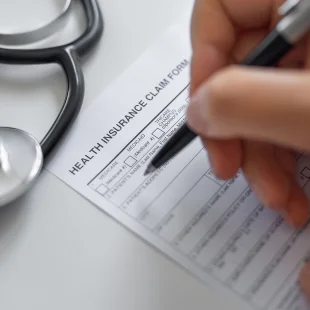 A person fills out a health insurance claim form with a pen. A stethoscope rests on the table beside the form.