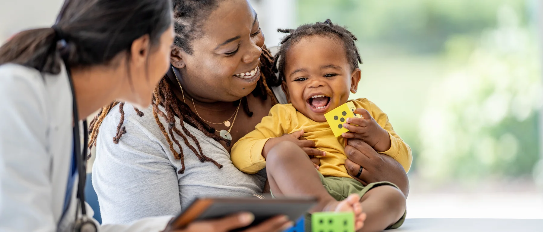 A doctor holding a tablet looking at a smiling mother holding her laughing toddler, who is playing with colorful toy blocks during a check-up.
