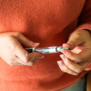 A person wearing an orange sweater holds an insulin pen, preparing to use it by removing the green cap to manage their insulin levels.