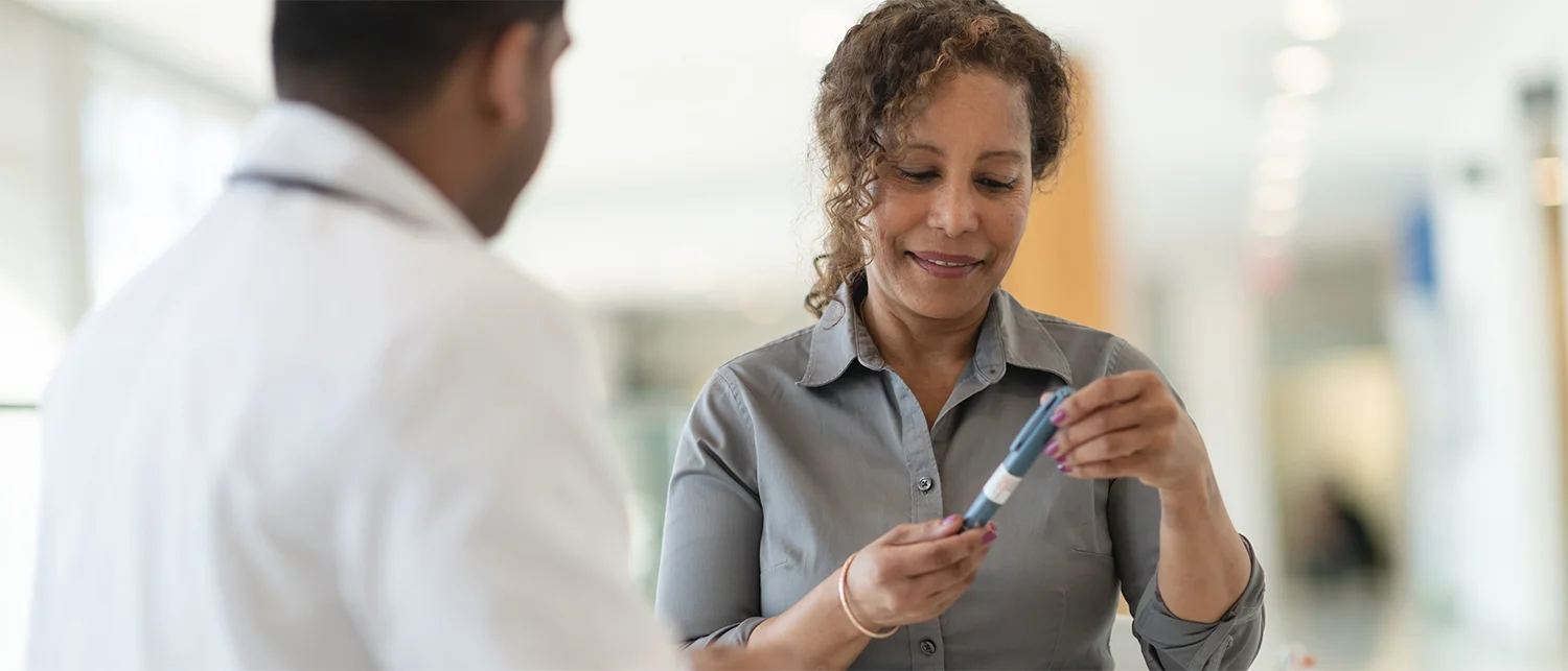 A woman smiles while holding an insulin pen, sitting across from a doctor in a white coat in a bright, modern medical setting. Insulin supplies are neatly arranged on the table.