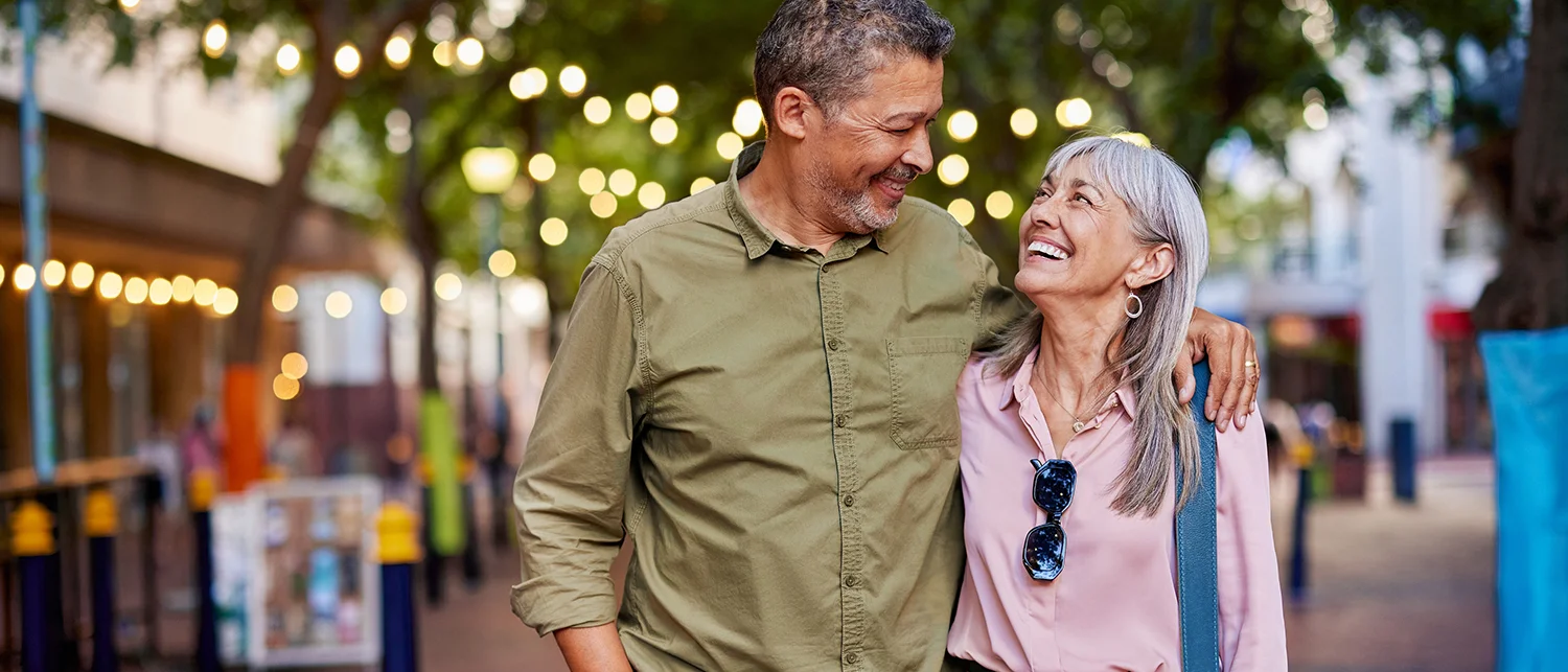 An older couple smiles at each other while walking outdoors under string lights discussing Medicare options.
