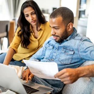 A young couple sit together on a couch, looking at a laptop and holding paperwork. They appear focused on the health insurance forms.