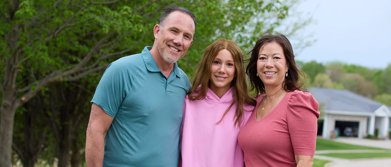 Three people stand closely together outdoors, facing the camera. A person in a teal shirt, a person in a pink hoodie, and a person in a coral top pose in front of green trees.