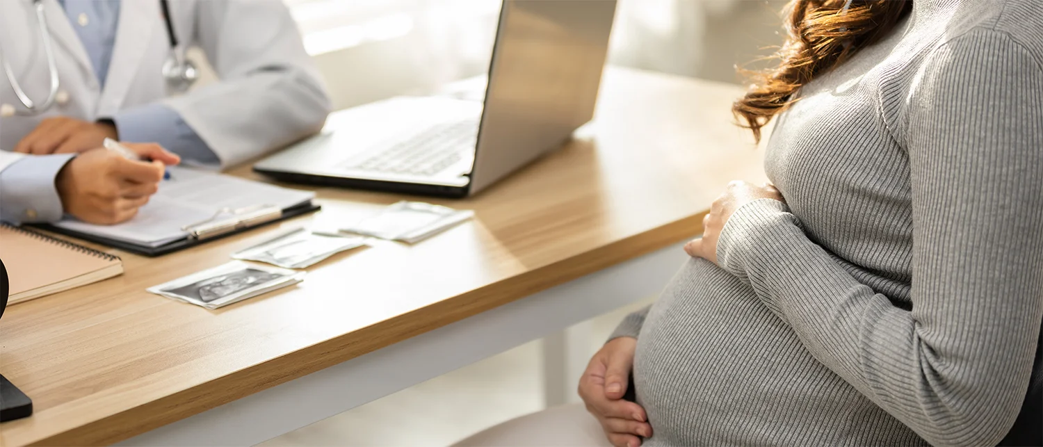 A pregnant person sits across from a doctor at a desk with ultrasound images, a notebook, and a laptop during a medical consultation.