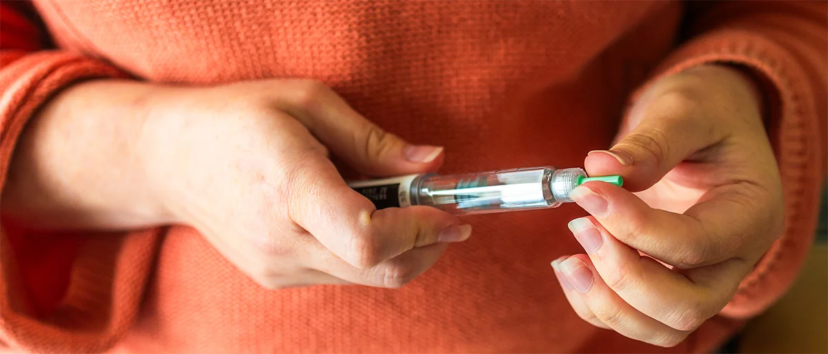 A person wearing an orange sweater holds an insulin pen, preparing to use it by removing the green cap to manage their insulin levels.