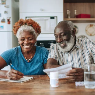 An older couple sit at a kitchen table, smiling while reviewing papers together; the woman writes notes, and a glass of water and medicine bottle are nearby.