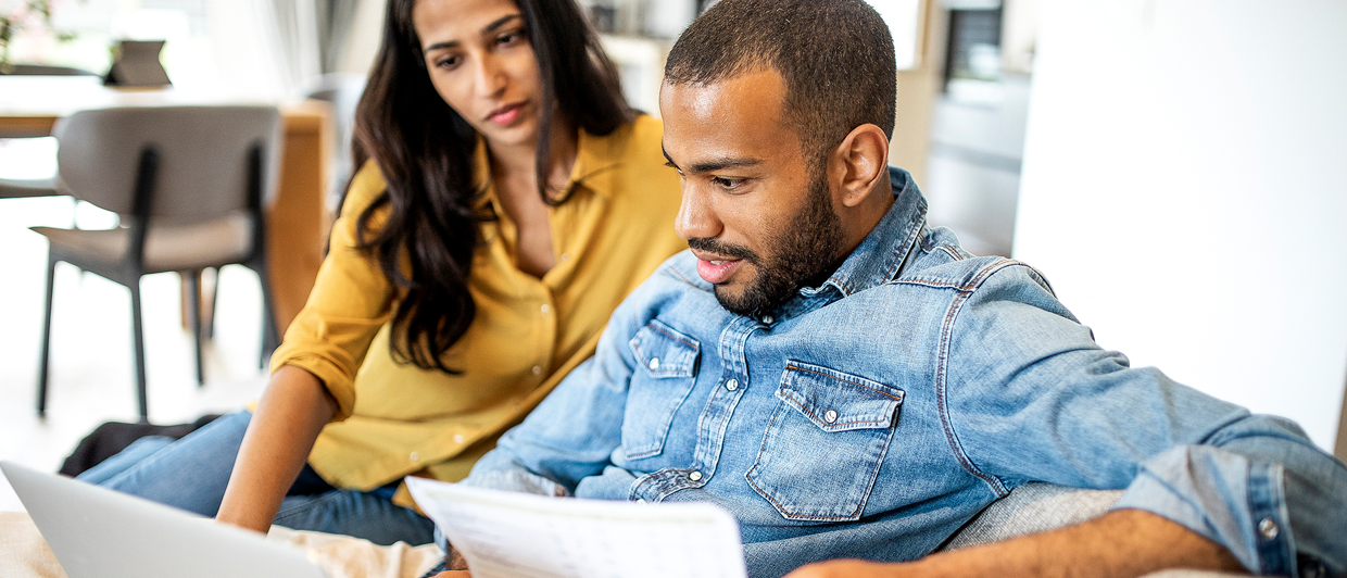 A young couple sit together on a couch, looking at a laptop and holding paperwork. They appear focused on the health insurance forms.