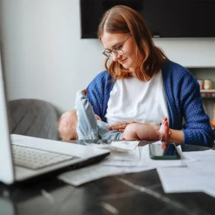 A woman wearing glasses looking at the baby on her lap while sitting at a desk with a laptop.