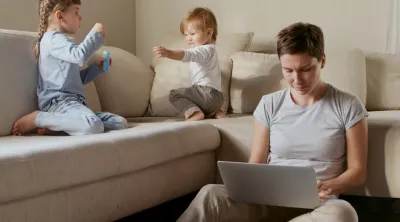 A woman is on laptop with two children blowing bubbles on couch next to her.
