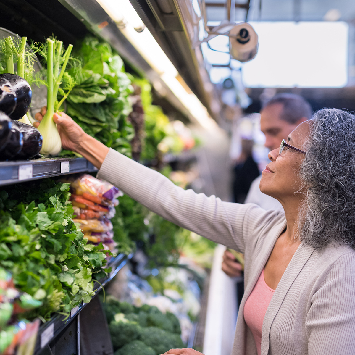 An older woman with glasses selects fresh fennel from a produce shelf in a grocery store, surrounded by leafy greens and vegetables.
